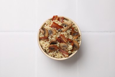 Photo of Tasty quinoa porridge with fried bacon and mushrooms in bowl on white tiled table, top view