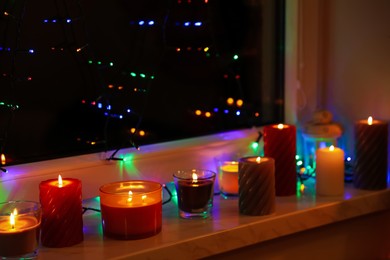 Photo of Many burning candles and festive lights on window sill indoors. Cosy atmosphere