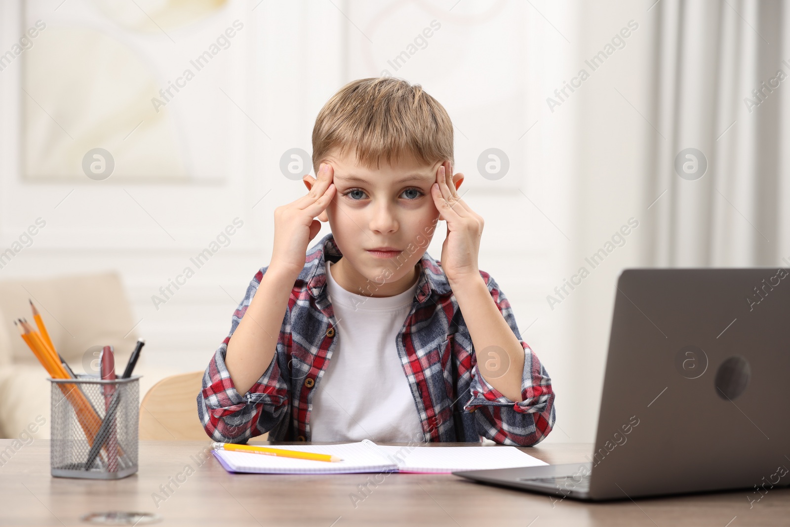 Photo of Little boy suffering from headache at wooden desk indoors