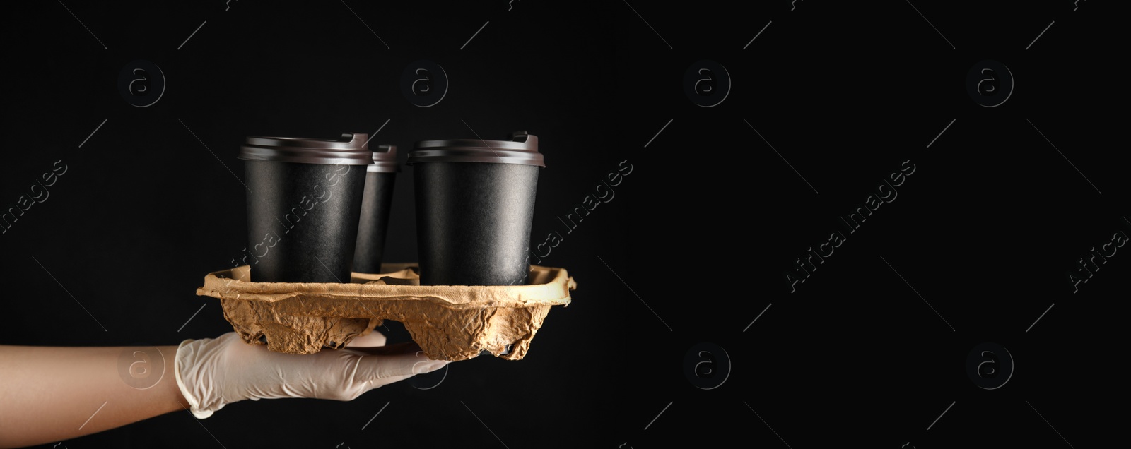 Image of Woman holding cardboard holder with takeaway paper coffee cups on black background, closeup. Banner design with space for text