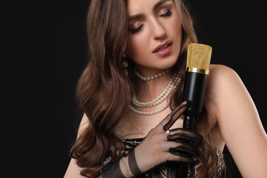 Beautiful young woman in stylish dress with microphone singing on black background