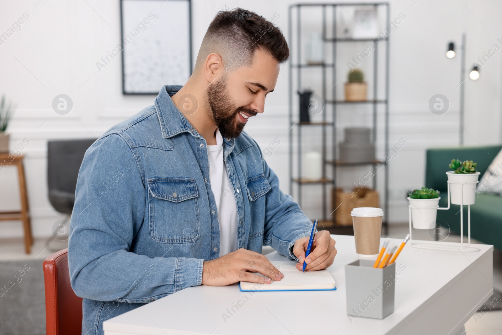 Photo of Young man writing in notebook at white table indoors