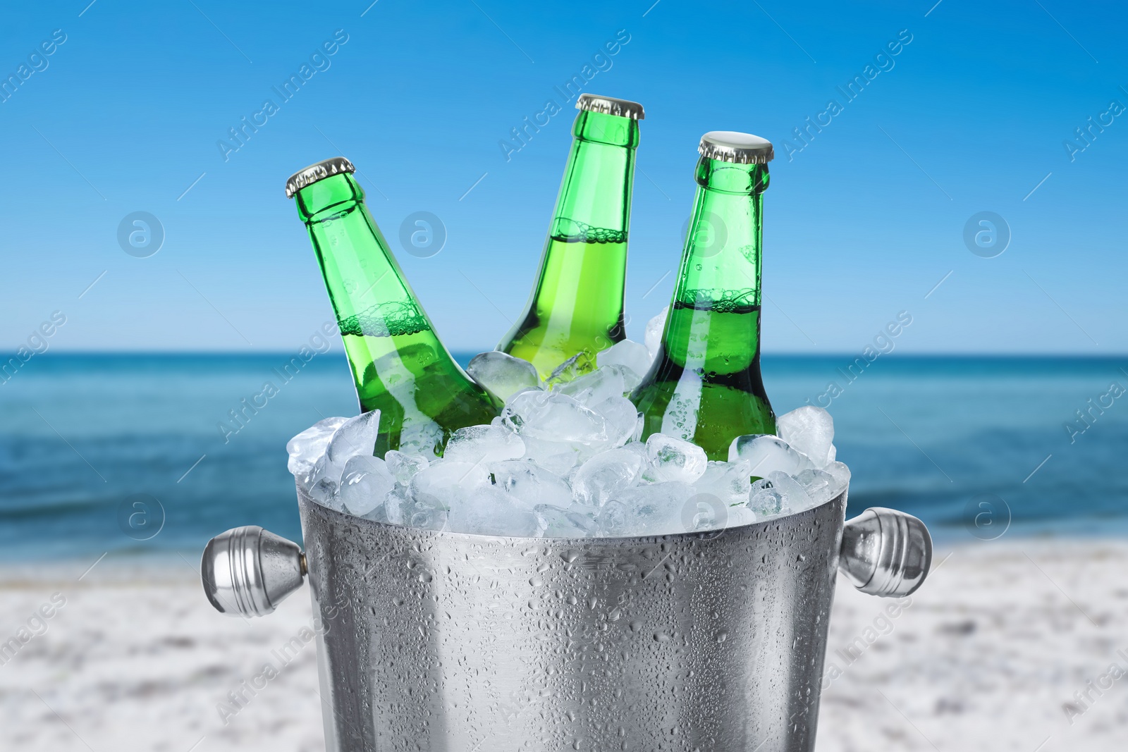 Image of Bottles of beer with ice cubes in metal bucket against blurred sea and sandy beach