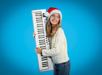 Photo of Young woman in Santa hat with synthesizer on light blue background. Christmas music