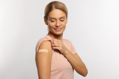 Smiling woman with adhesive bandage on arm after vaccination on light background