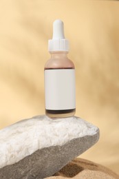 Photo of Bottle of serum and stone on sand against beige background