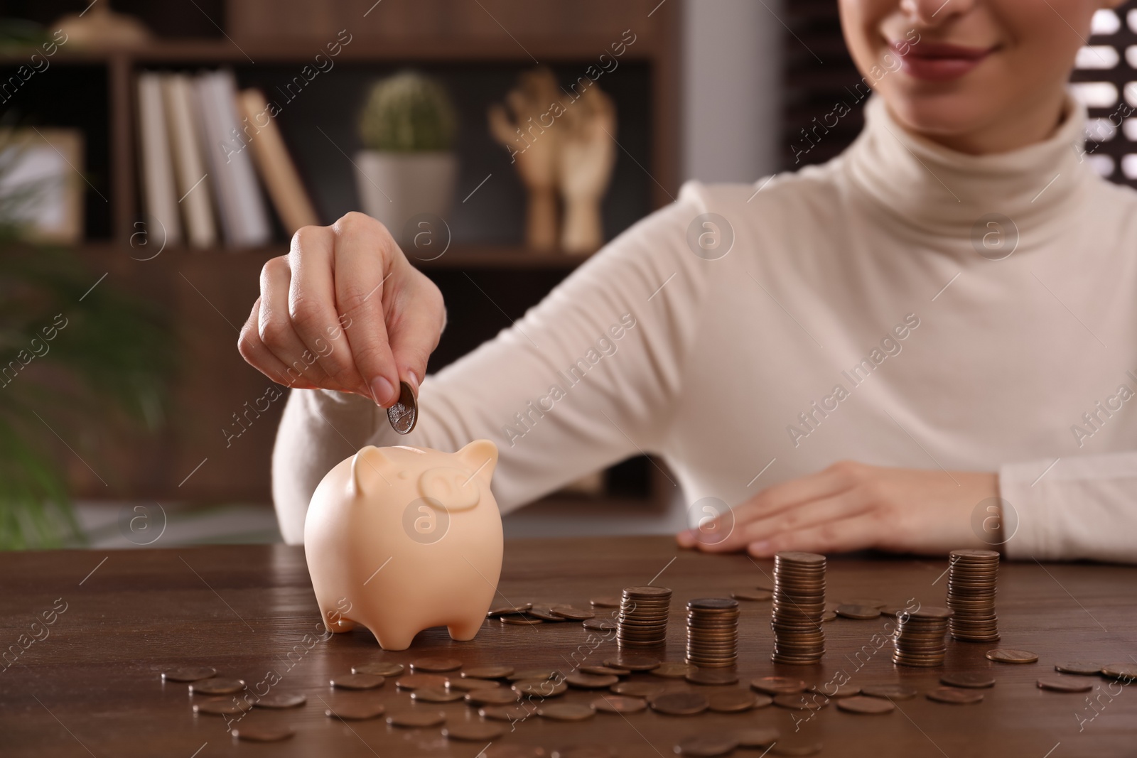 Photo of Woman putting money into piggy bank at wooden table indoors, closeup