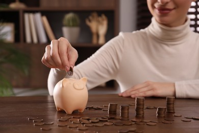 Photo of Woman putting money into piggy bank at wooden table indoors, closeup