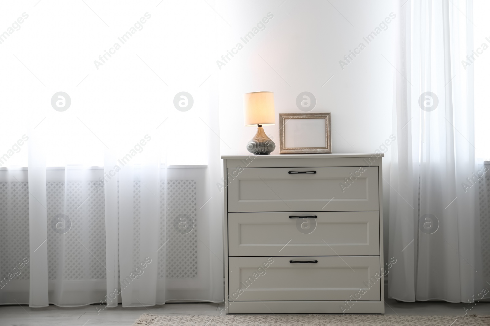 Photo of Modern chest of drawers with lamp near window in room