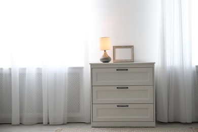 Photo of Modern chest of drawers with lamp near window in room