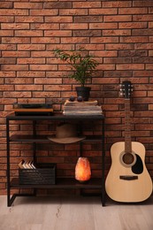 Photo of Stylish turntable, guitar and vinyl records on shelving unit near red brick wall indoors