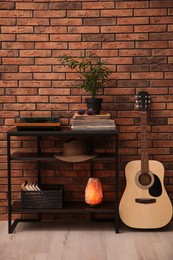 Stylish turntable, guitar and vinyl records on shelving unit near red brick wall indoors