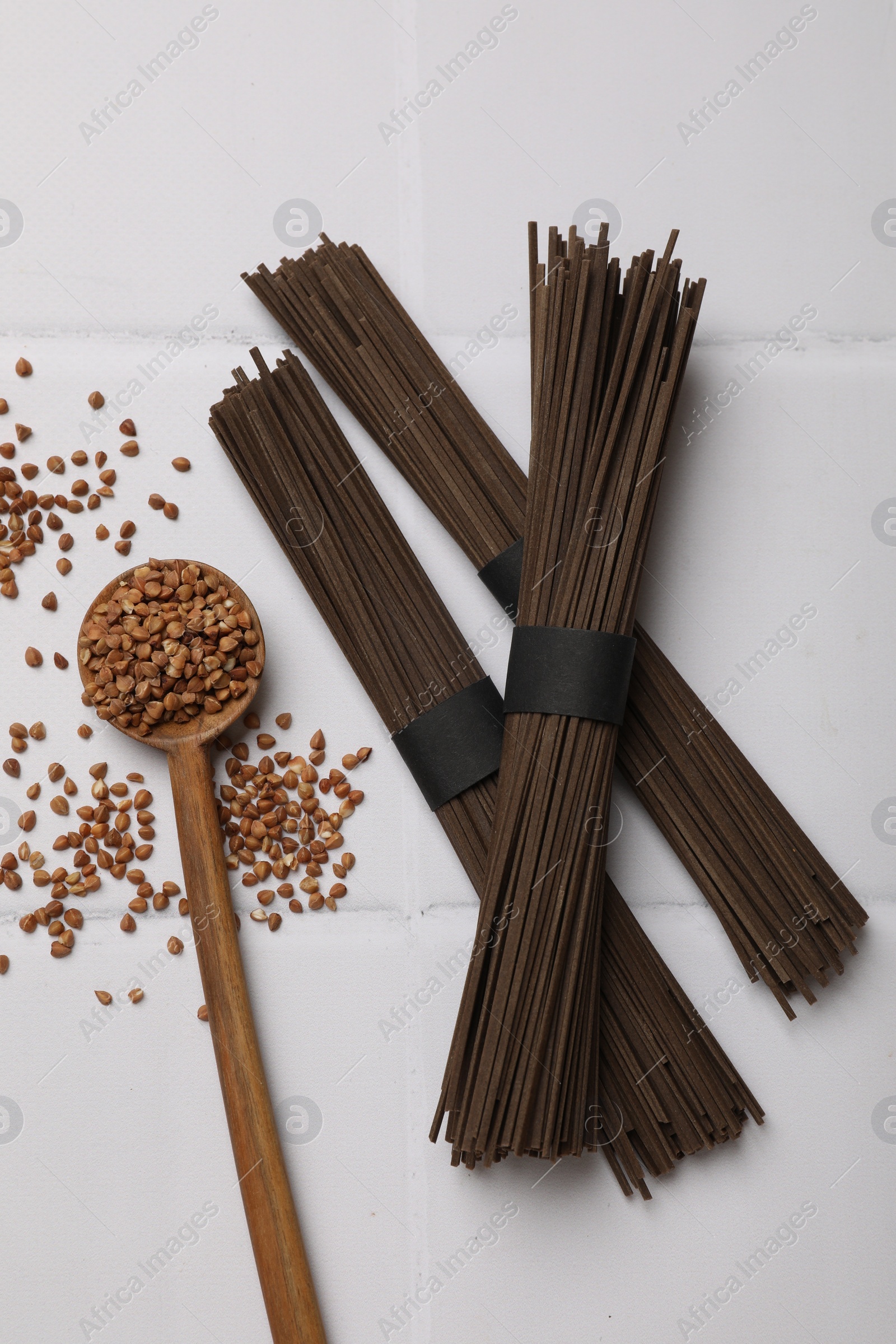 Photo of Uncooked buckwheat noodles (soba), grains and spoon on white tiled table, flat lay
