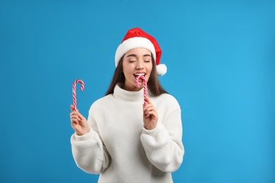 Photo of Young woman in beige sweater and Santa hat holding candy canes on blue background. Celebrating Christmas