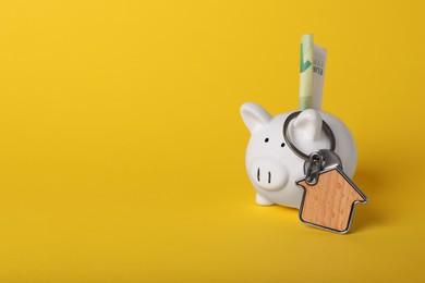 Ceramic piggy bank with euro banknote and key trinket on yellow background, space for text. Financial savings