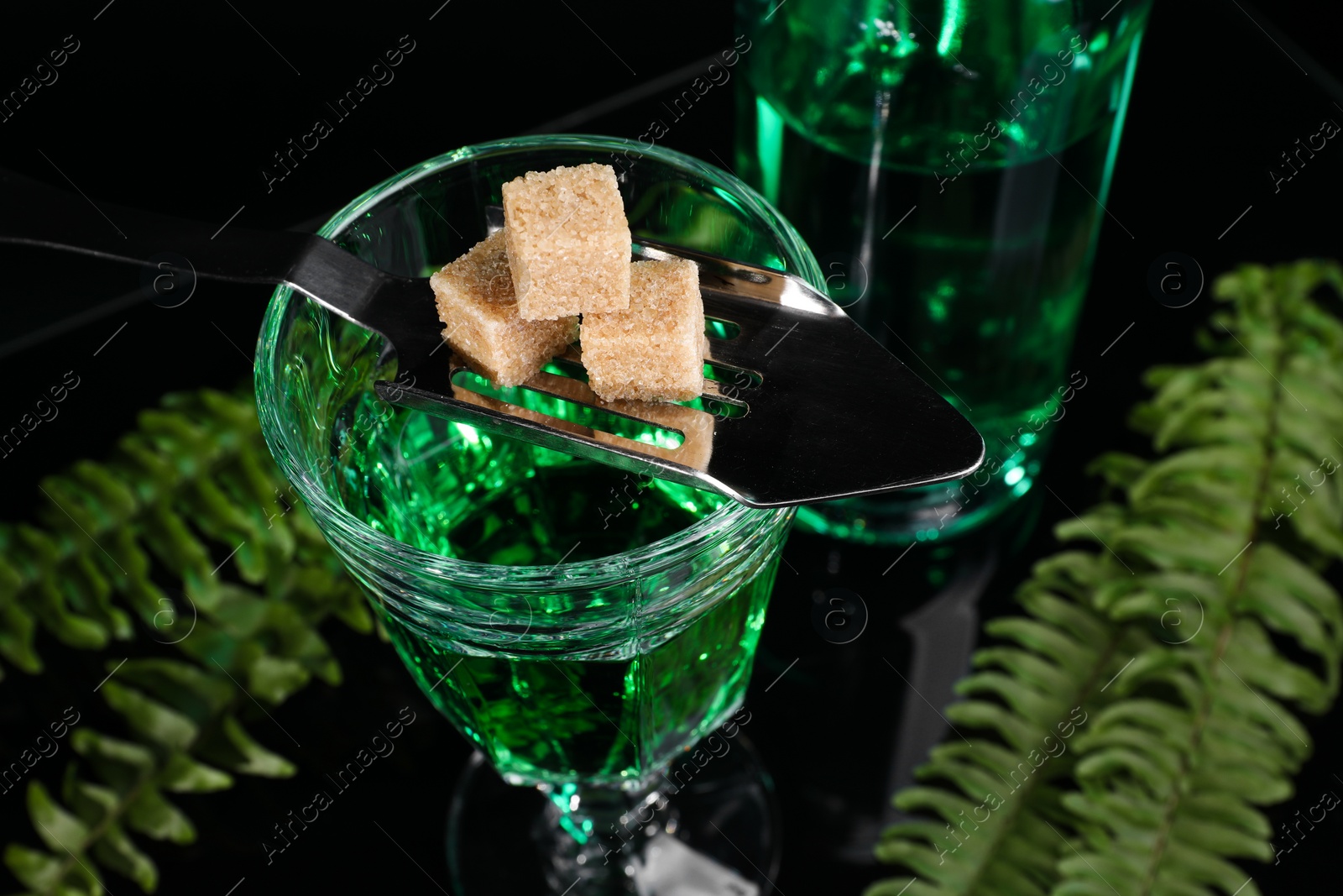 Photo of Absinthe in glass, brown sugar, spoon and green leaves on mirror table, closeup. Alcoholic drink