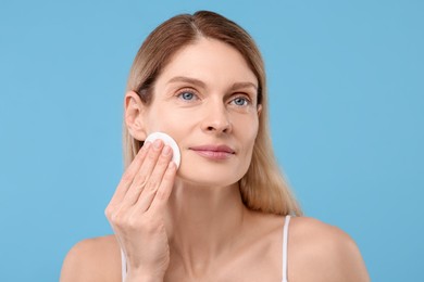 Beautiful woman removing makeup with cotton pad on light blue background