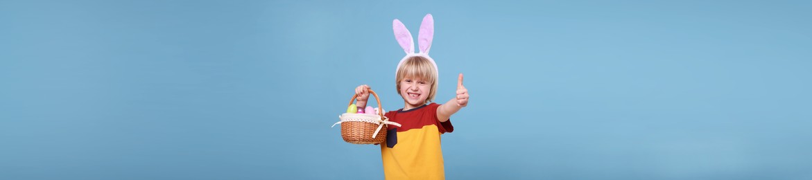 Happy boy with bunny ears holding basket full of Easter eggs and showing thumbs up on light blue background. Banner design