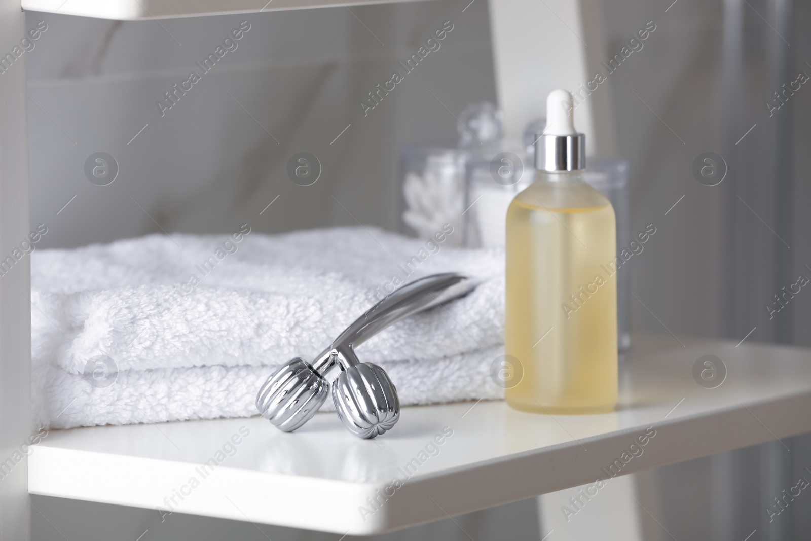 Photo of Metal face roller, towels and serum on shelving unit in bathroom