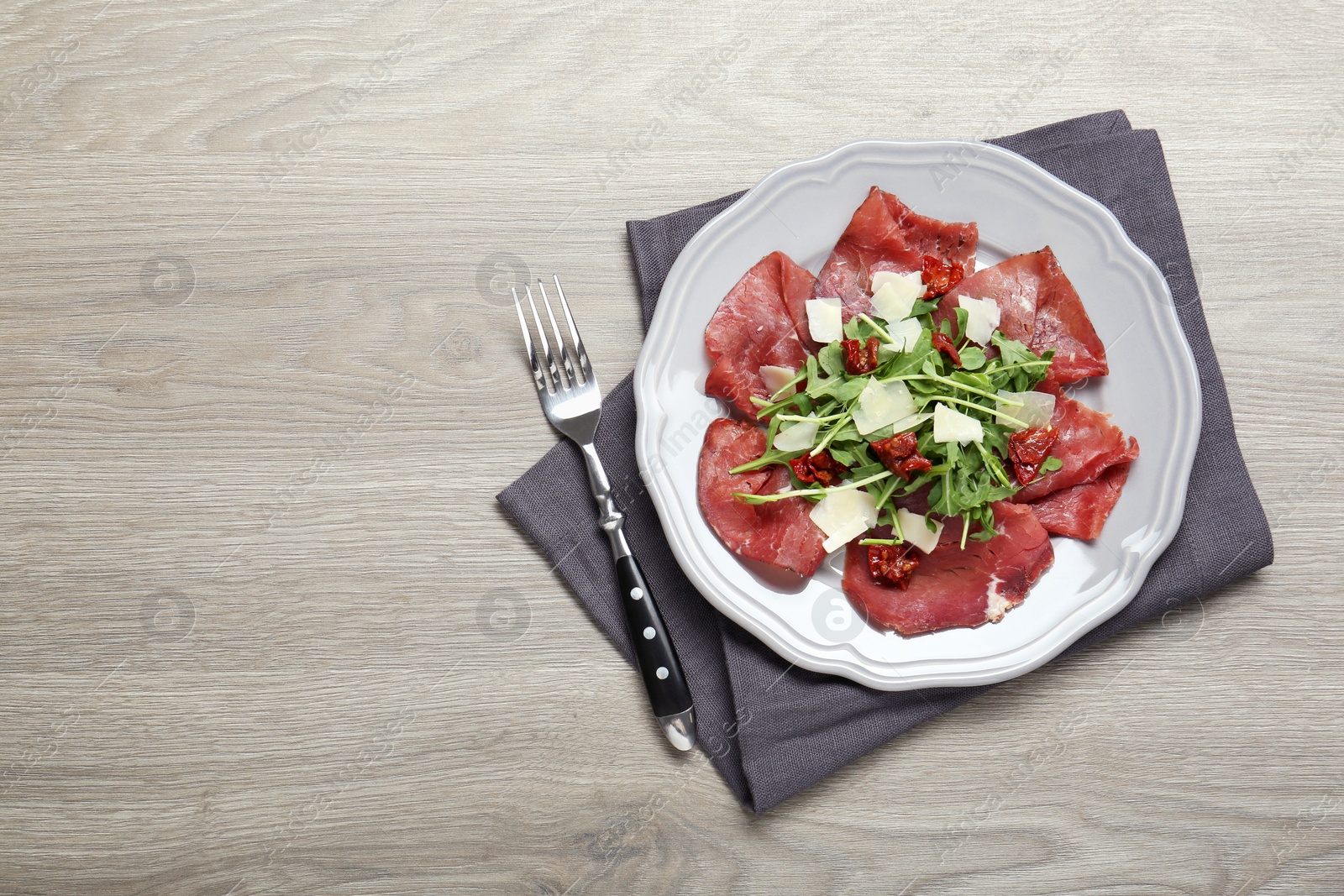 Photo of Plate of tasty bresaola salad with sun-dried tomatoes, parmesan cheese and fork on wooden table, top view. Space for text