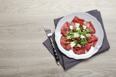 Plate of tasty bresaola salad with sun-dried tomatoes, parmesan cheese and fork on wooden table, top view. Space for text