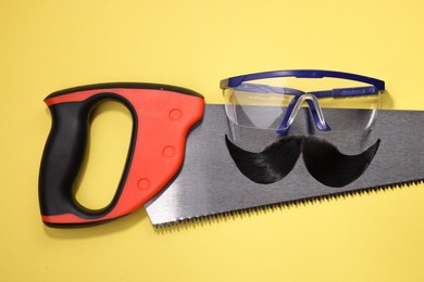 Man's face made of artificial mustache, safety glasses and hand saw on yellow background, top view