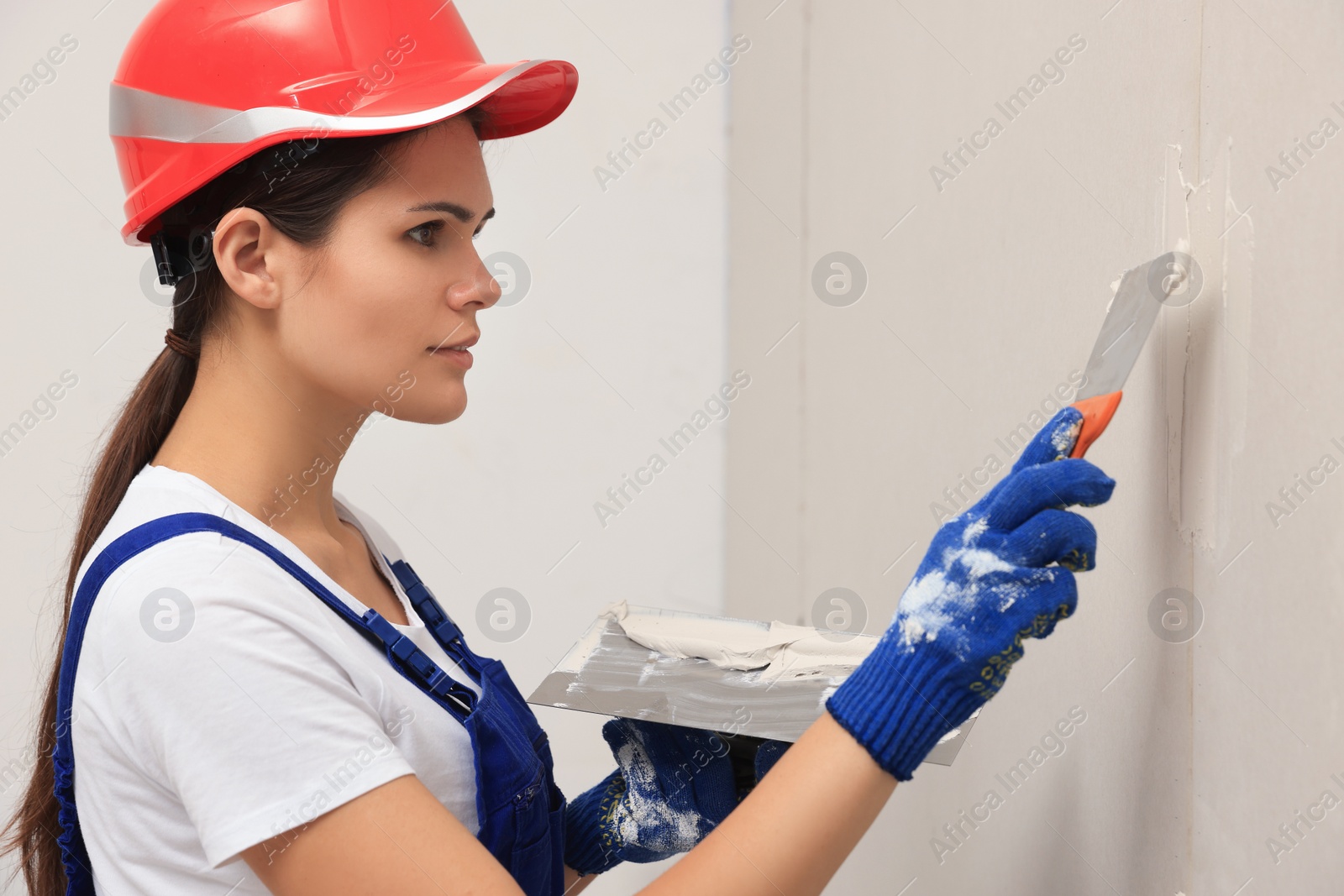 Photo of Professional worker in hard hat plastering wall with putty knives indoors
