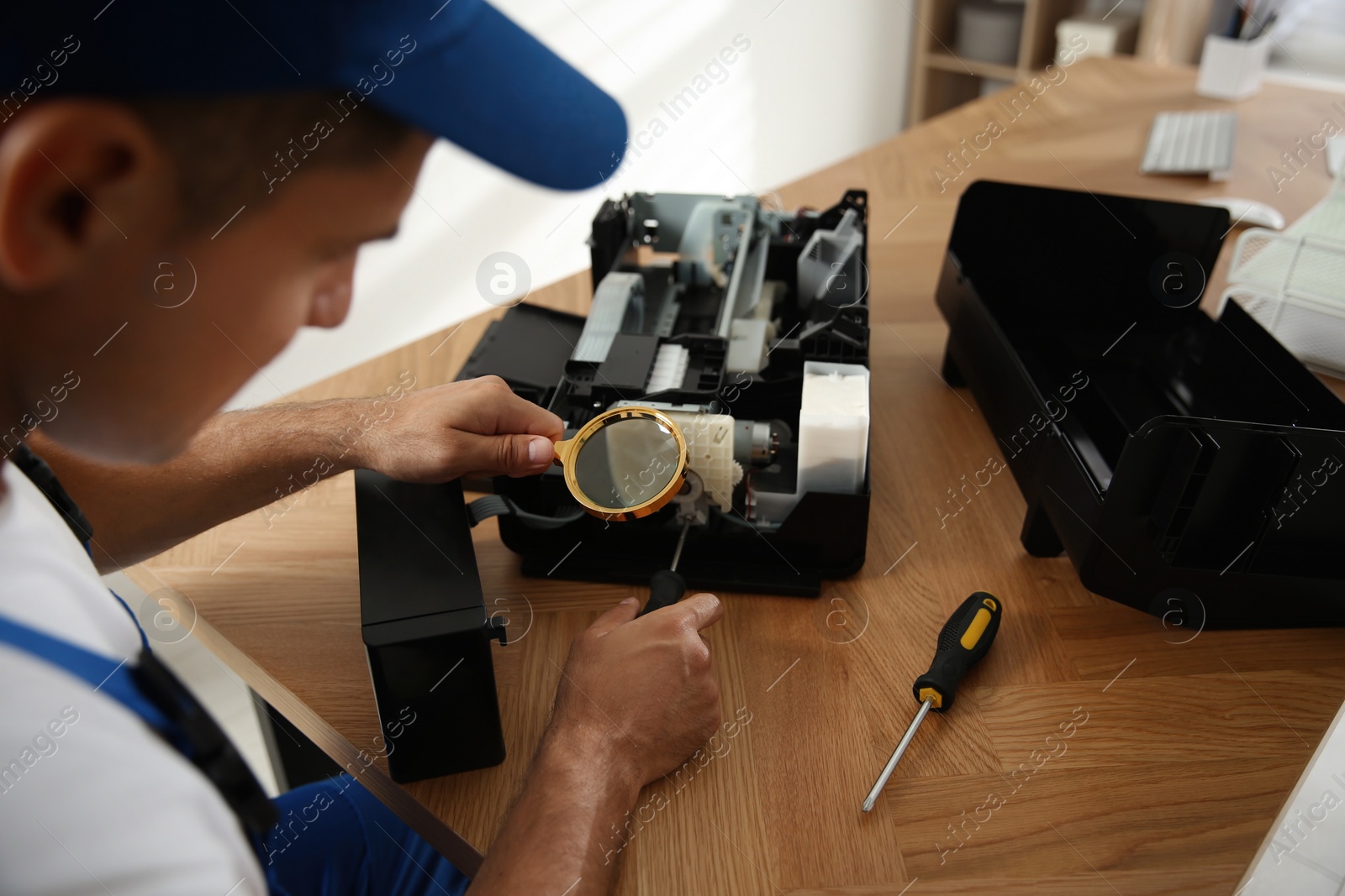 Photo of Repairman with magnifying glass and screwdriver fixing modern printer indoors, closeup