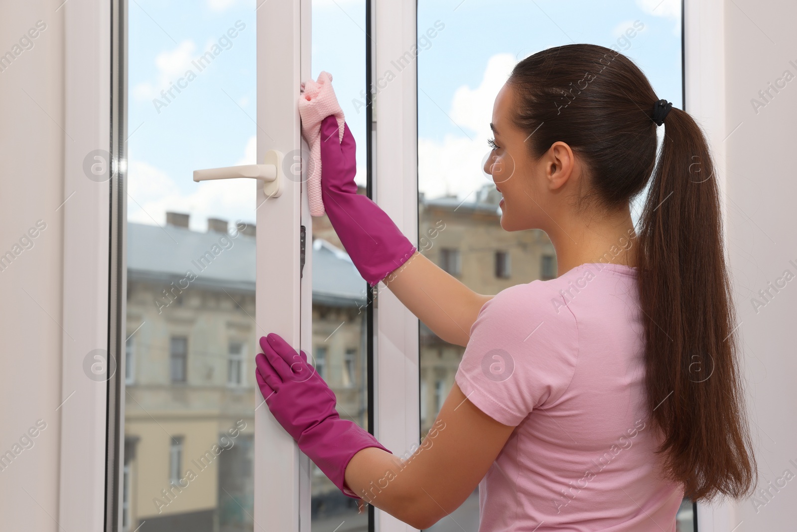 Photo of Happy young woman cleaning window frame with rag at home