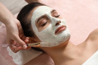 Cosmetologist applying white mask onto woman's face in spa salon, closeup