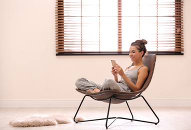 Photo of Young woman using smartphone near window with blinds at home. Space for text