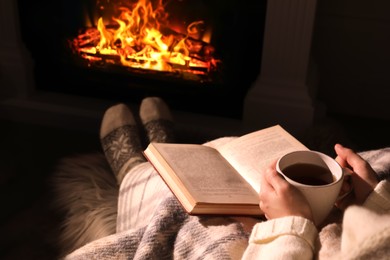Woman with book and cup near fireplace indoors, closeup. Cozy atmosphere