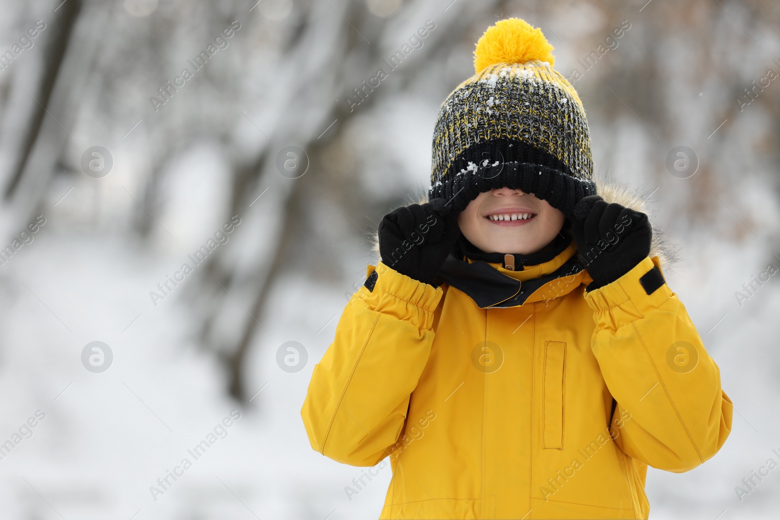 Photo of Cute little boy covering eyes with hat in snowy park on winter day, space for text