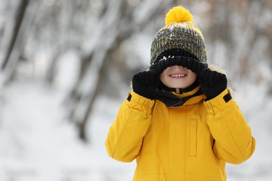 Cute little boy covering eyes with hat in snowy park on winter day, space for text