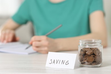 Financial savings. Woman making notes at white wooden table indoors, focus on glass jar with coins