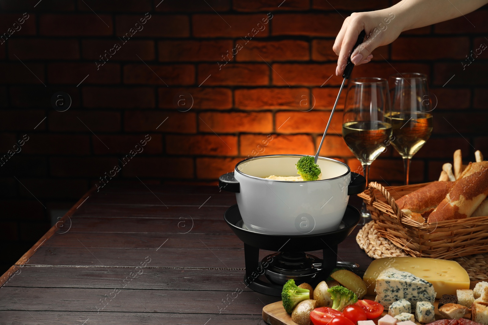 Photo of Woman dipping piece of broccoli into fondue pot with melted cheese at wooden table with wine and snacks, closeup. Space for text