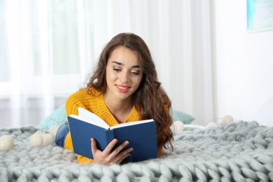 Young beautiful woman in warm sweater reading book on bed at home