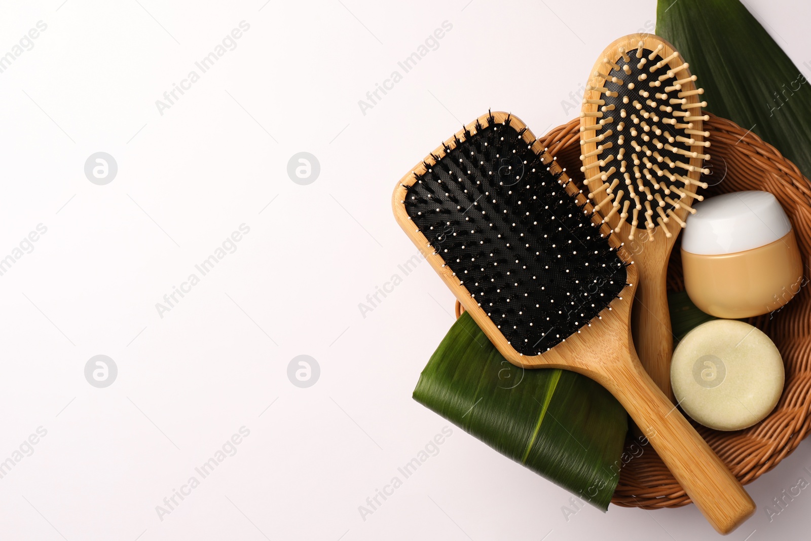Photo of Wicker basket with wooden hairbrushes, cosmetic products and green leaves on white background, top view. Space for text