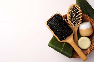 Wicker basket with wooden hairbrushes, cosmetic products and green leaves on white background, top view. Space for text