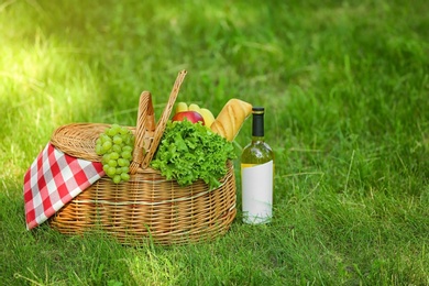 Photo of Wicker basket with blanket, wine and food on green grass in park, space for text. Summer picnic