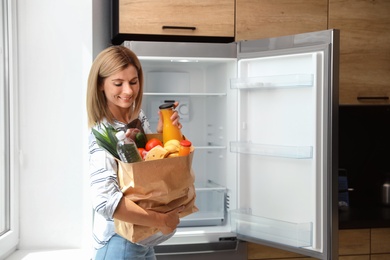 Woman with paper bag full of products near refrigerator in kitchen