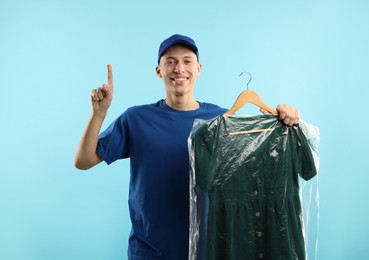 Dry-cleaning delivery. Happy courier holding dress in plastic bag and pointing at something on light blue background, space for text