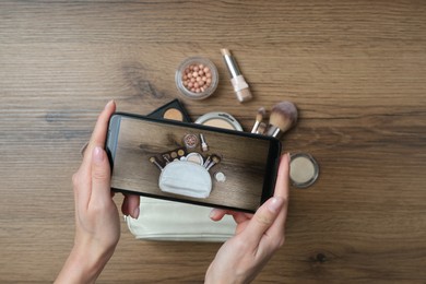 Photo of Woman taking photo of makeup products with smartphone at wooden table, top view