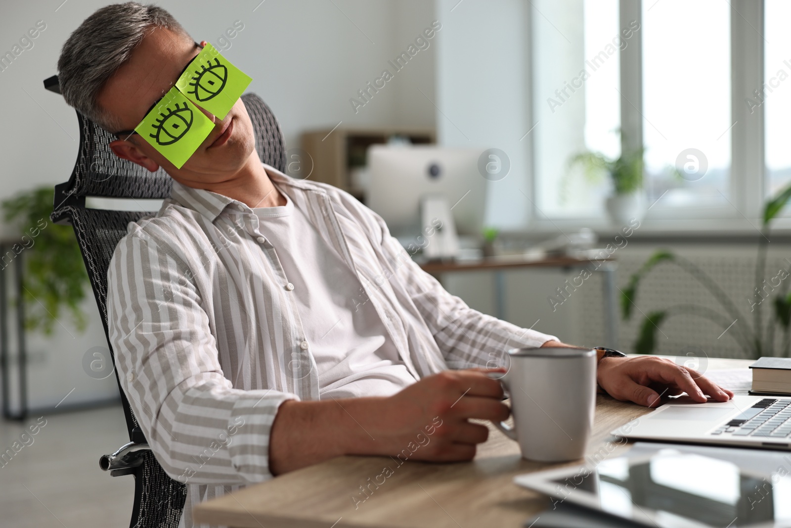 Photo of Man with fake eyes painted on sticky notes snoozing at workplace in office