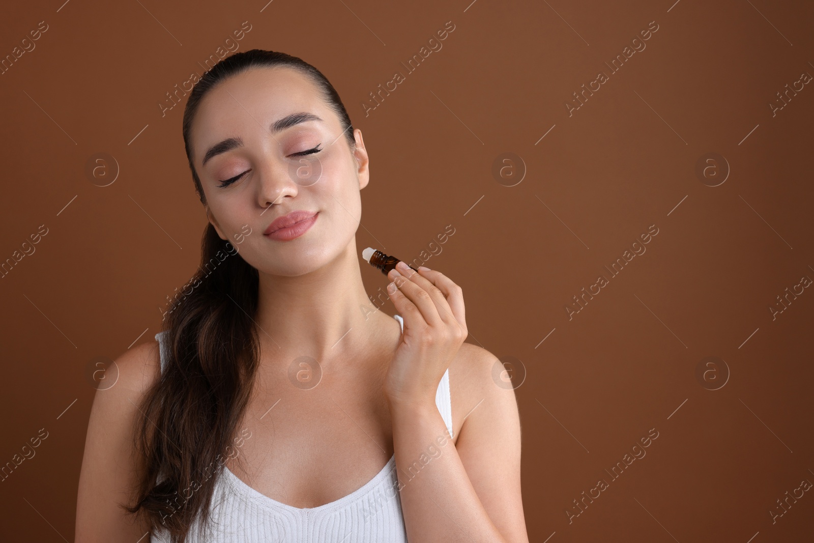 Photo of Beautiful young woman with roller bottle applying essential oil onto neck on brown background, space for text
