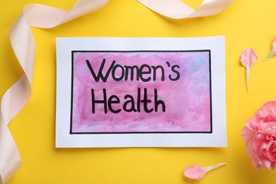 Sheet of paper with words Women's Health, ribbon and carnation flower on yellow background, flat lay