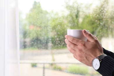 Young man with cup of coffee near window indoors on rainy day, closeup