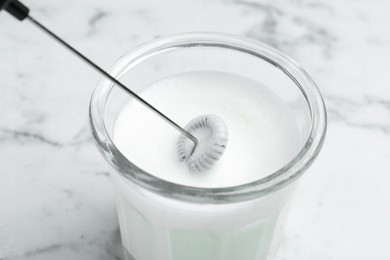 Whisking milk in glass with mini mixer (milk frother) at white marble table, closeup