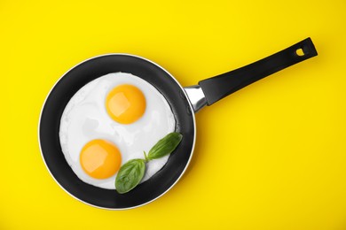 Tasty fried eggs with basil in pan on yellow background, top view