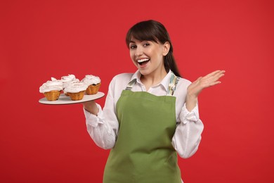 Happy professional confectioner in apron holding delicious cupcakes on red background